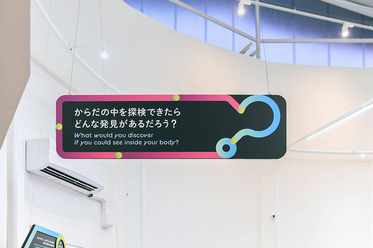 宙吊りの黒いパネルに「からだの中を探検できたら？」と未来への問いを示す展示サイン。来場者の想像力を刺激する仕掛けを象徴するデザイン。