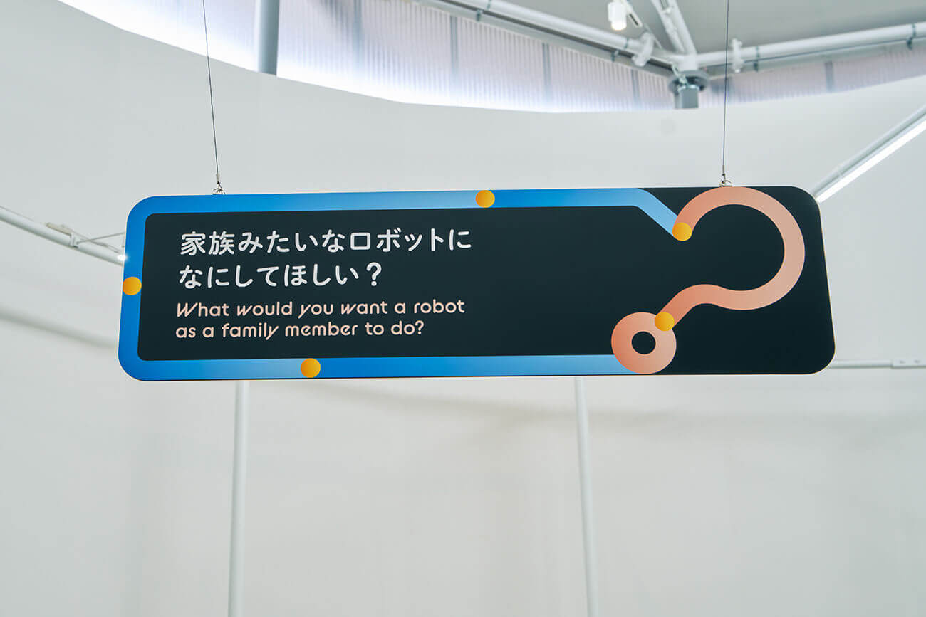 空中に掲げられた青縁のパネルに「家族みたいなロボットに何をしてほしい？」と問いが示され、来場者の想像を促す展示サイン。