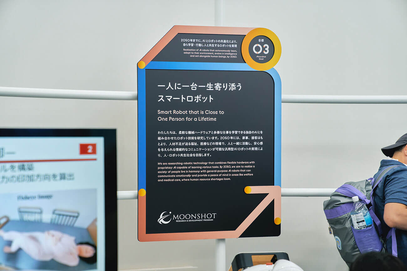 ムーンショット目標3を紹介する縦型パネルが立ち、一人に寄り添うスマートロボットの研究概要が日本語と英語で示されている展示説明板。