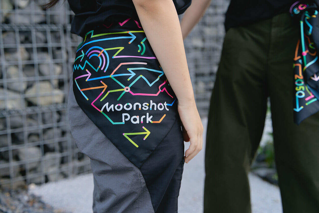 スタッフが腰に巻いた黒地のスカーフに、カラフルな矢印と「Moonshot Park」のロゴが描かれており、展示全体のビジュアル統一を示すアイテムとして写っている。