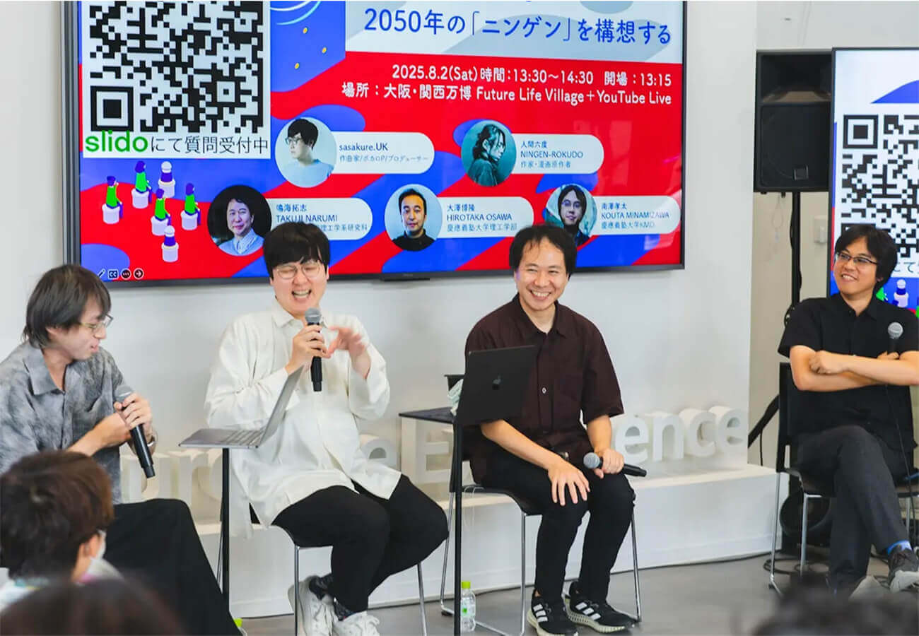 研究者やクリエイターが壇上でマイクを手に議論し、来場者が聞き入るステージイベントの様子。背景スクリーンには「2050年のニンゲンを構想する」企画の案内が表示されている。