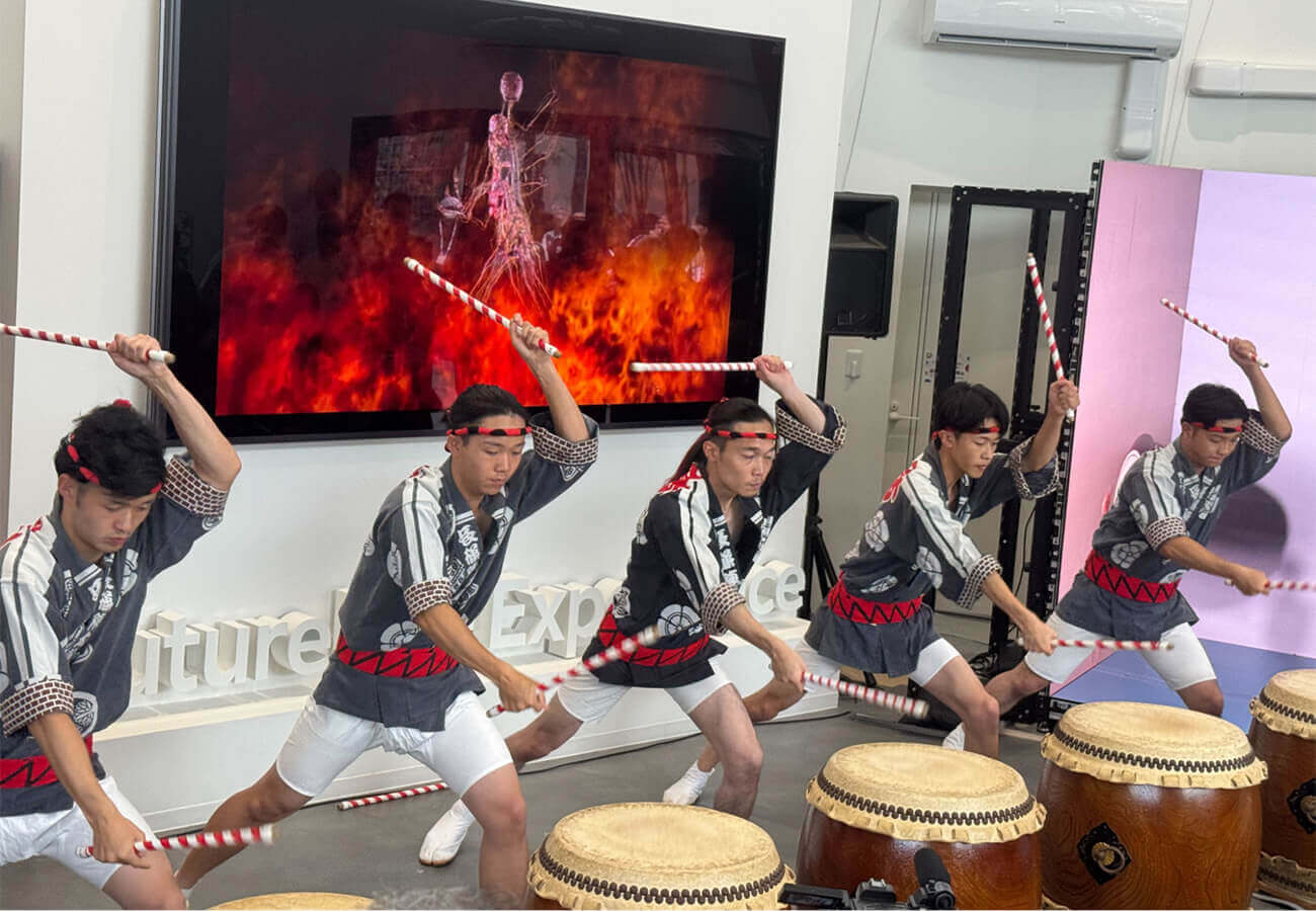和太鼓奏者の若者たちが隊列を組んで力強く演奏し、背後の大型モニターには炎の映像が映る。展示イベント内のライブパフォーマンスの様子。