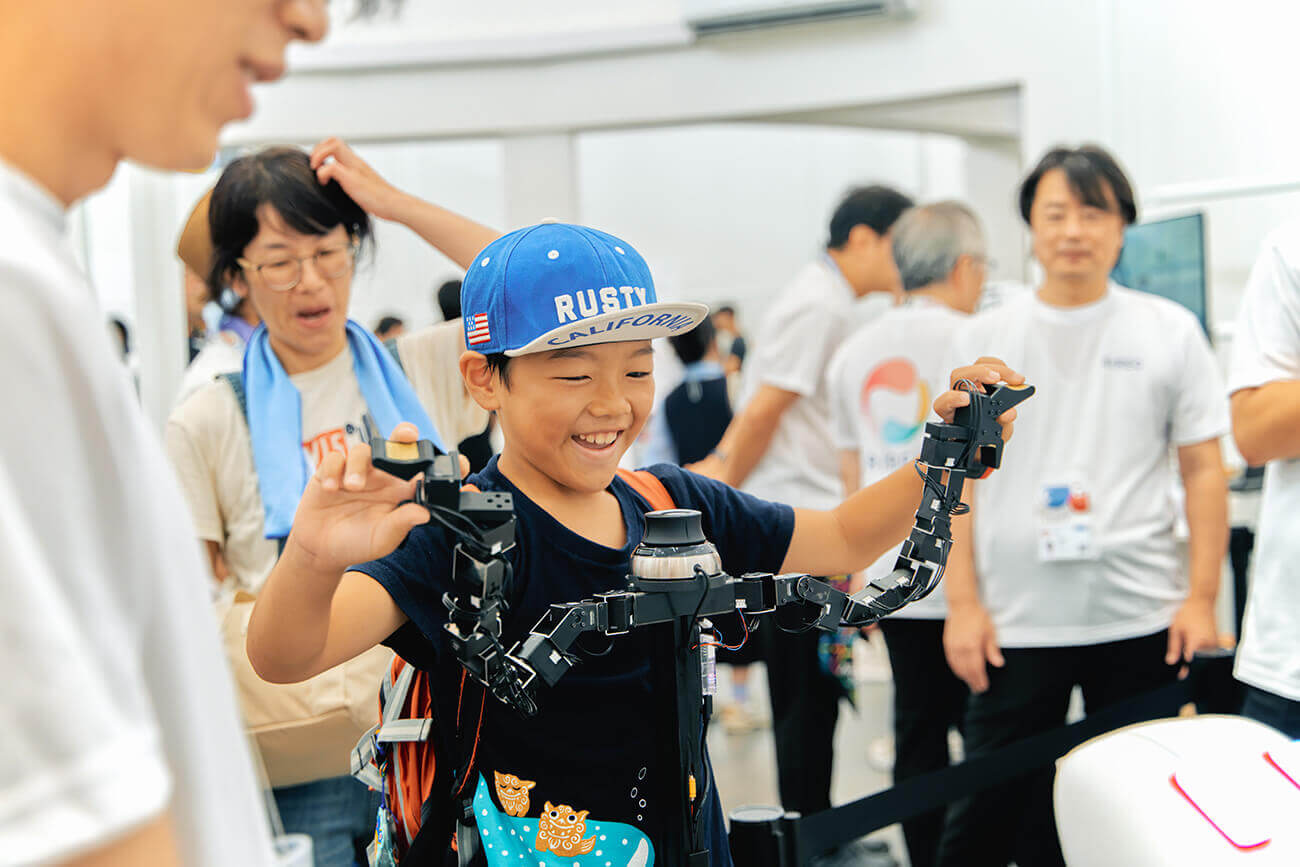 帽子をかぶった男の子が多関節ロボットアームを操作し、笑顔で動作を試している。周囲では大人たちが見守り、来場者が先端技術を体験する展示の一場面。