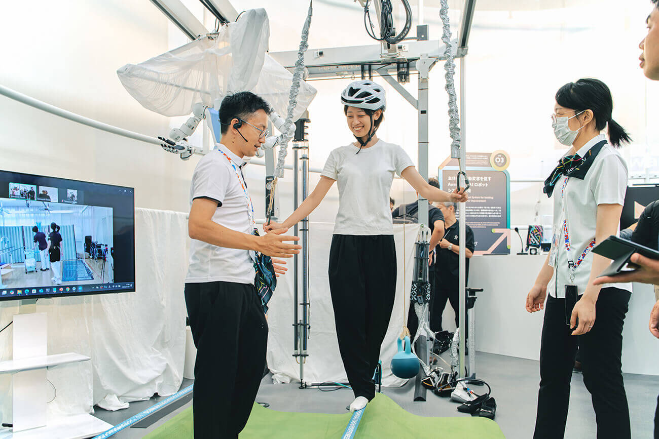 ヘルメットを着けた来場者が歩行支援装置の上でバランスを取り、スタッフが横で見守りながら操作を説明している様子。研究技術を実際に試せる体験展示。