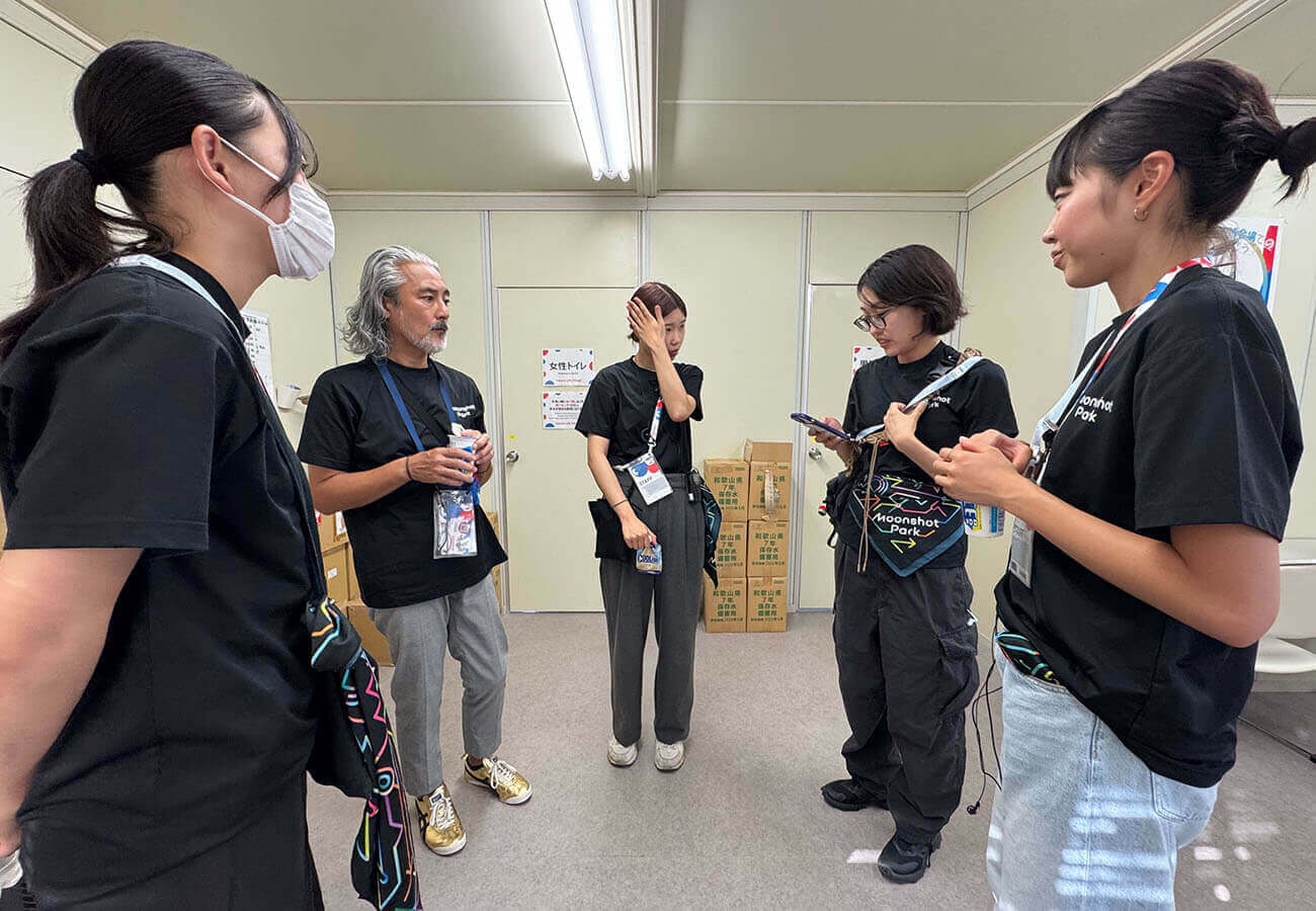 黒いスタッフTシャツを着たメンバーが控室で立ち話をし、担当内容の確認や共有を行っている様子。展示運営前のオリエンテーションの場面。