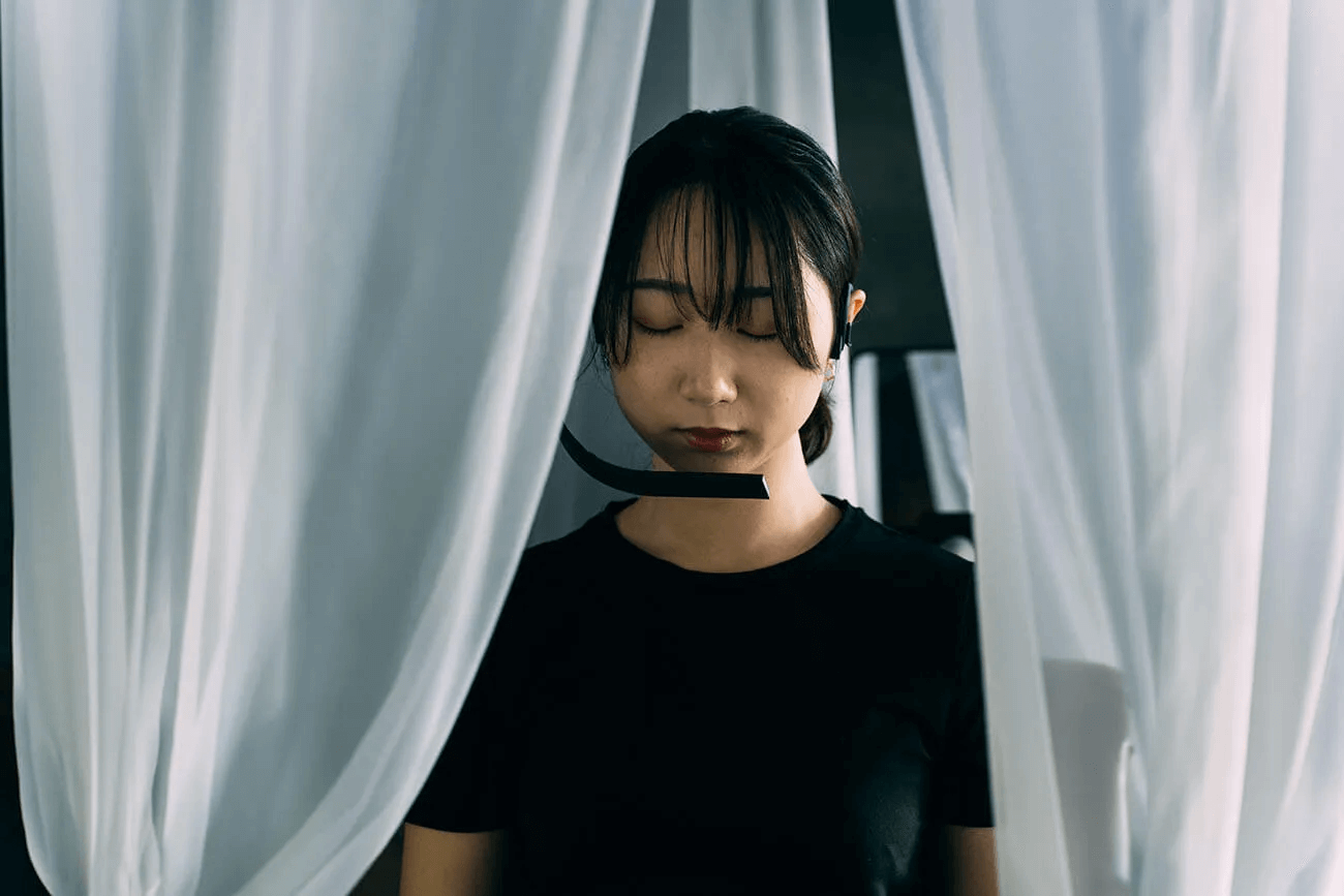 写真：佐野さん制作の作品の展示風景。女性が自身の呼吸を計測するデバイスの前でたたずみ、自身の呼吸のリズムや深さを骨伝導デバイスから感知している