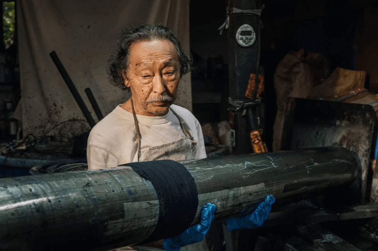 嵐絞りの復元作家・早川嘉英さんが、布を巻き付けた長い木製の棒を抱え、染料槽の脇で作業を見定める。薄暗い工房でエプロン姿の熟練職人が真剣な表情を浮かべる瞬間。