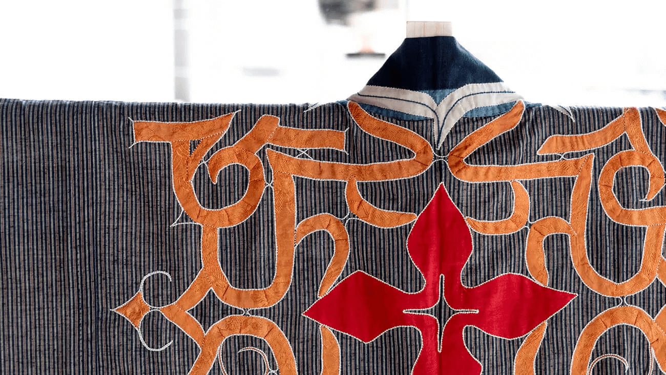 アイヌの民族衣装。灰色の布地に、オレンジや朱色のアイヌ独特の紋様が縫い付けられている。