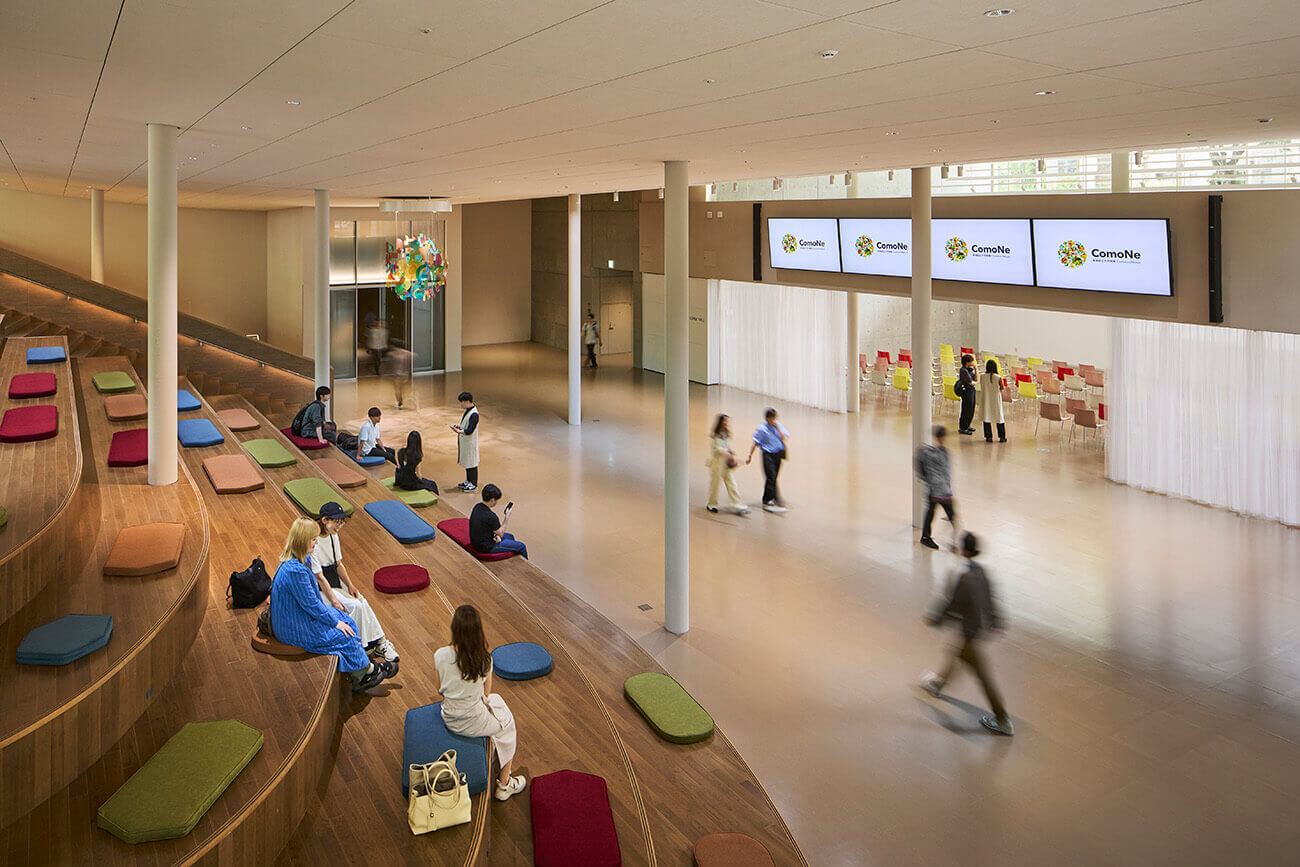 『ComoNe』施設内のラウンジスペース。床に座布団や低いテーブルが配置され、来館者がリラックスしたり談笑したりしながら過ごしている。吹き抜けから光が差し込む開放的な空間。