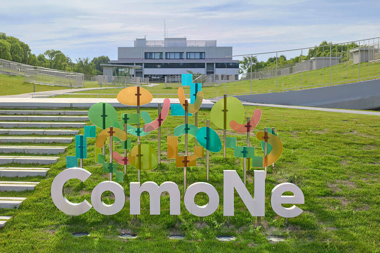 芝生広場に設置された屋外サイン。色とりどりの透明なパネルと立体文字が組み合わさり、背景には建物が見えている。