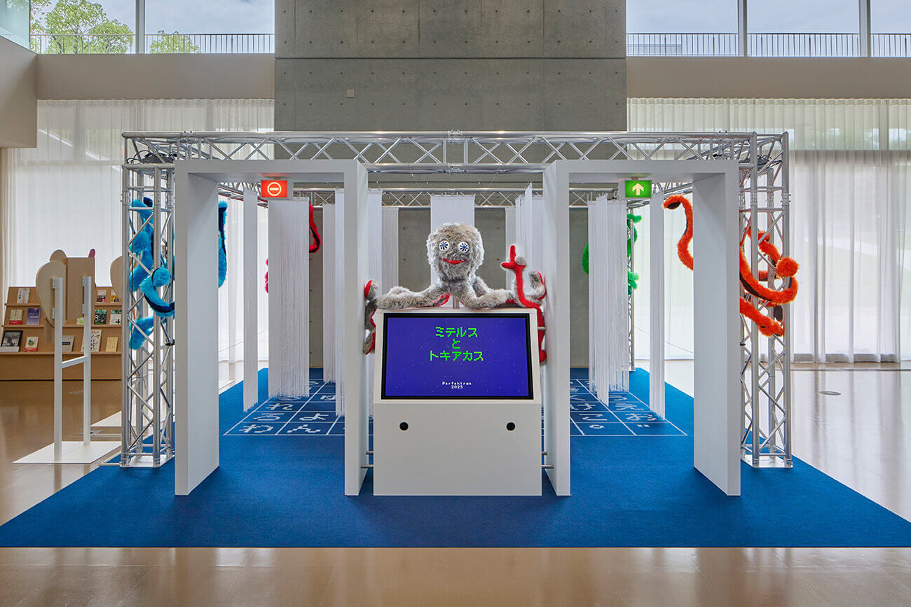 『ミテルスとトキアカス』の展示風景。中央にスクリーンとキャラクターが配置され、カラフルな装飾や布の仕切りが並ぶインスタレーション。