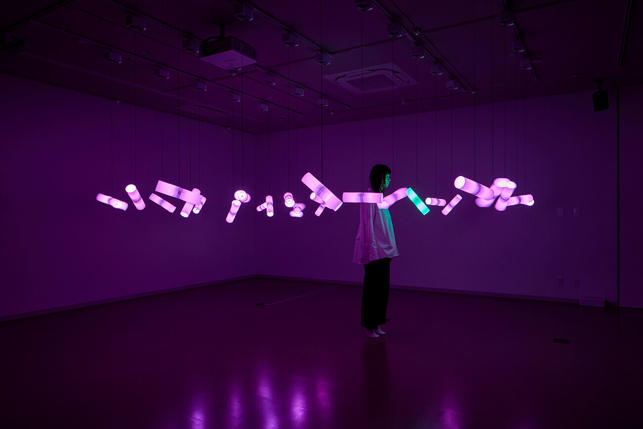 Fragmentations of Unityの展示。紫や緑に発光する円筒形のオブジェが吊り下げられているインスタレーション。