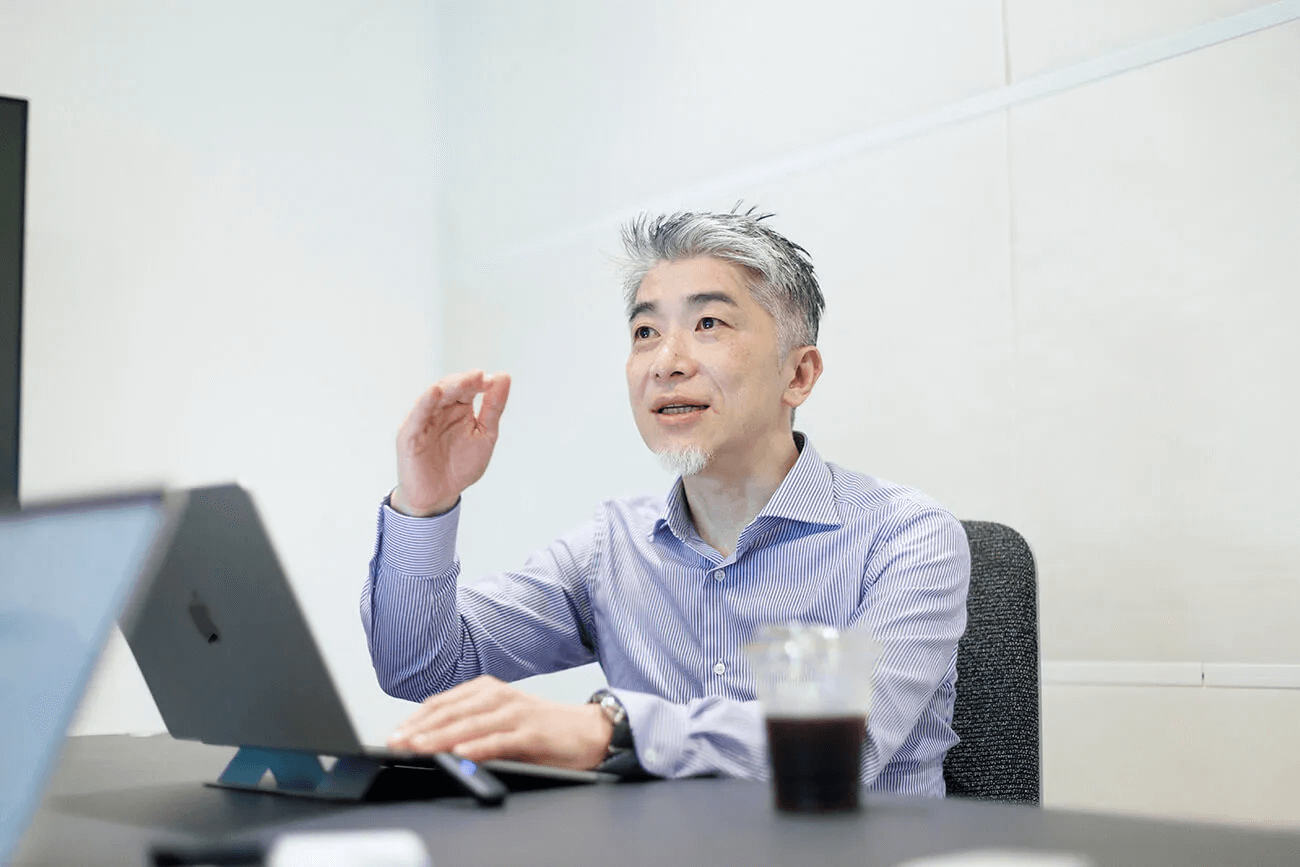 会議室でノートパソコンを前に、諏訪光洋が身振りを交えて説明している様子