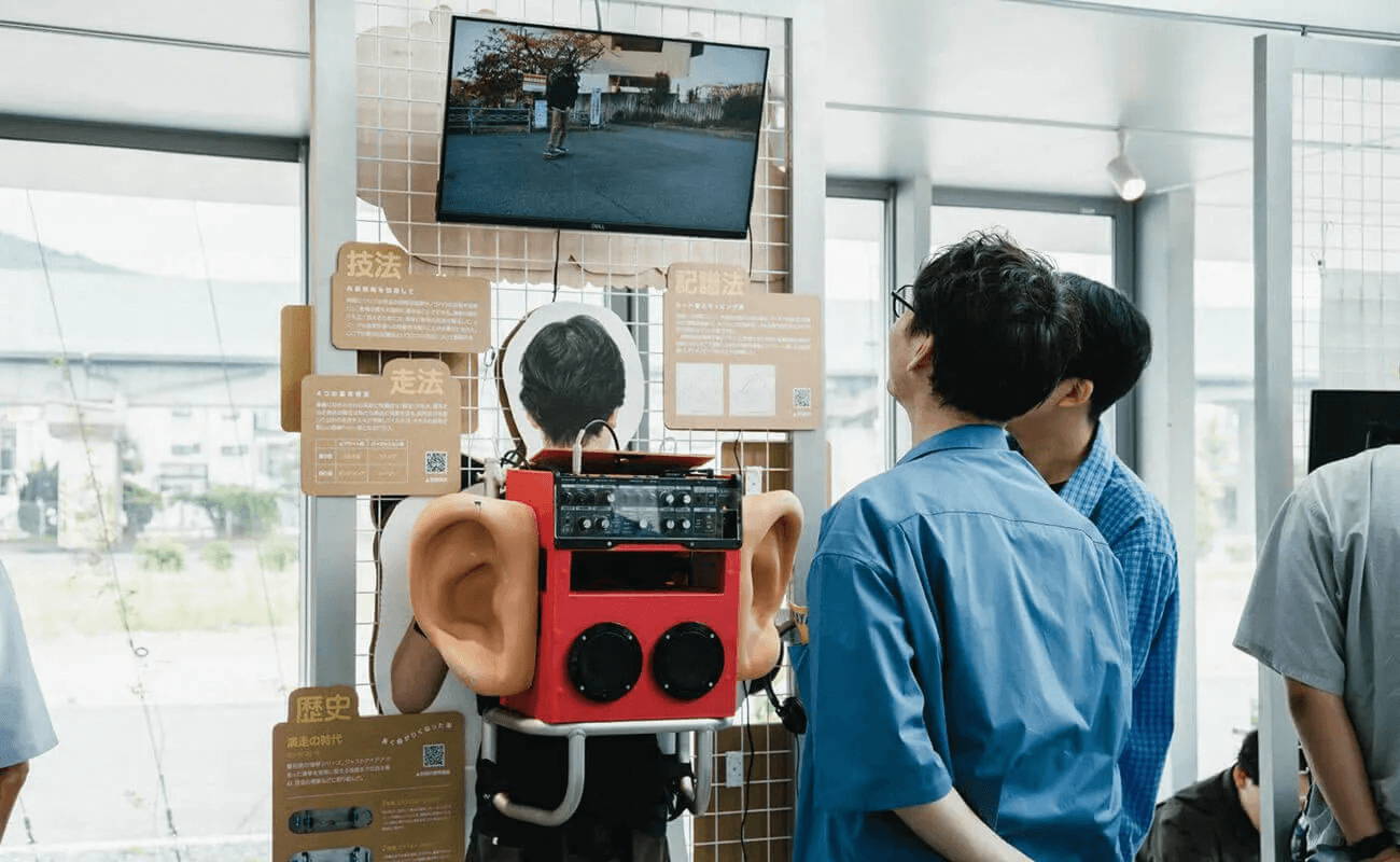 展示会場で、大きな耳の模型を取り付けた赤い装置の展示を来場者が覗き込み、上部のモニター映像を見ている
