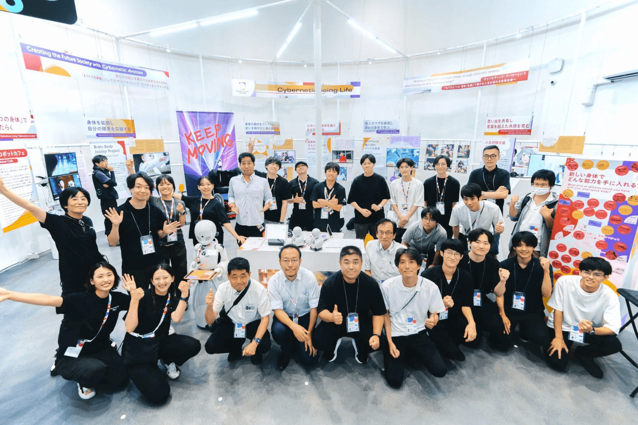 展示会場でスタッフや研究者たちが一堂に並び、笑顔でポーズを取る集合写真。体験展示を支えた多様なメンバーがそろい、達成感を味わっている雰囲気が伝わる。