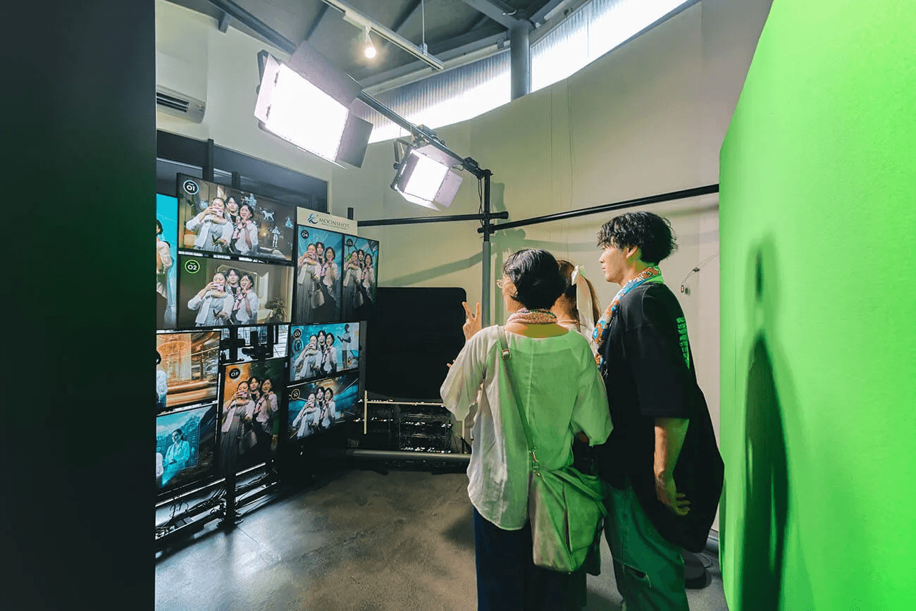 フォトスポットで、実際に参加者が記念撮影を行う様子。男女3名が、万博グッズを身纏いながら、一人がスマートフォンを構えて撮影を行なっている。三人の集合した様子が、10個のディスプレイにそれぞれ表示されている。