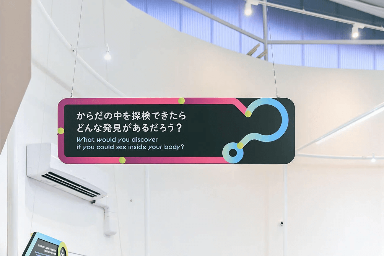 宙吊りの黒いパネルに「からだの中を探検できたら？」と未来への問いを示す展示サイン。来場者の想像力を刺激する仕掛けを象徴するデザイン。