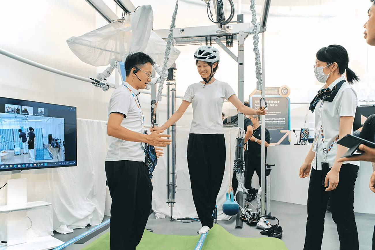 ヘルメットを着けた来場者が歩行支援装置の上でバランスを取り、スタッフが横で見守りながら操作を説明している様子。研究技術を実際に試せる体験展示。