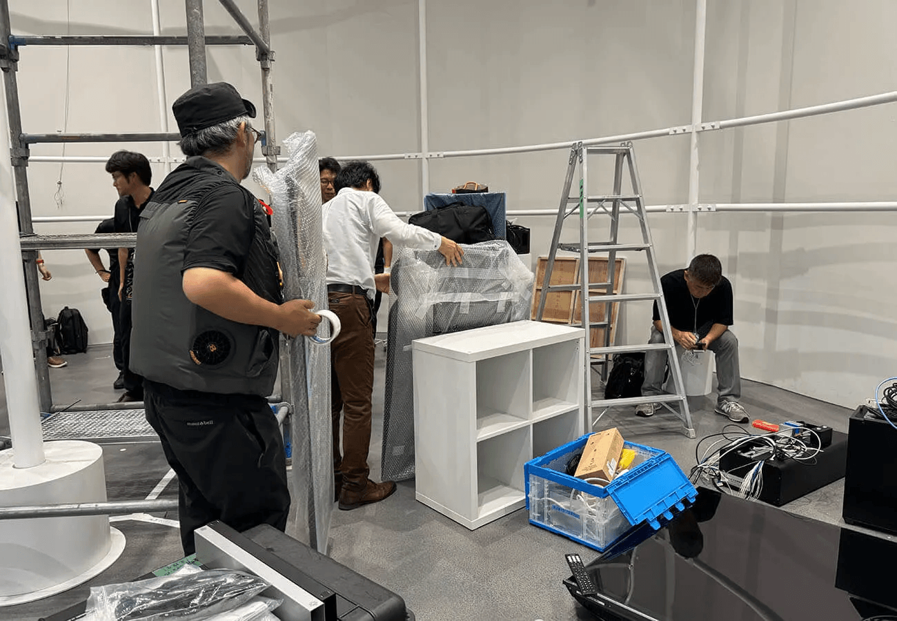 スタッフが棚や機材を梱包し、脚立や資材が並ぶ中で展示の準備を進めている設営作業の様子。週替わり展示の入れ替えを支える裏側を写す。