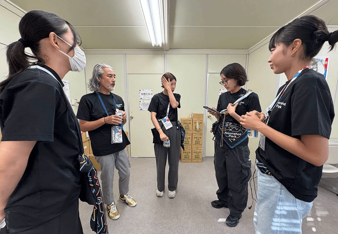 黒いスタッフTシャツを着たメンバーが控室で立ち話をし、担当内容の確認や共有を行っている様子。展示運営前のオリエンテーションの場面。