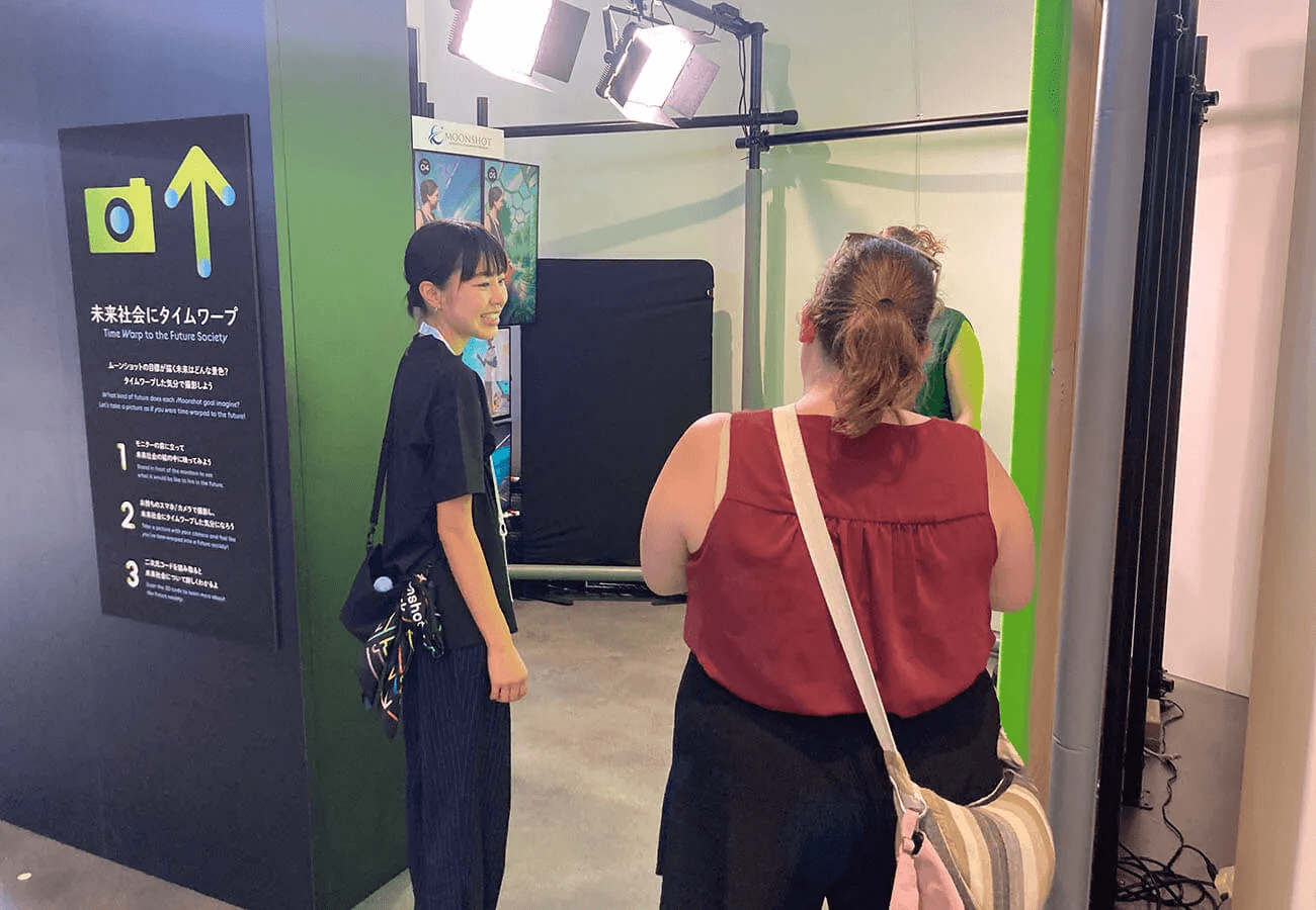 撮影ブースで海外からの来場者がスタッフの案内を受け、未来社会の背景に自分を投影する体験を準備している様子。ライトが照らす中、黒い案内パネルが設置されている。